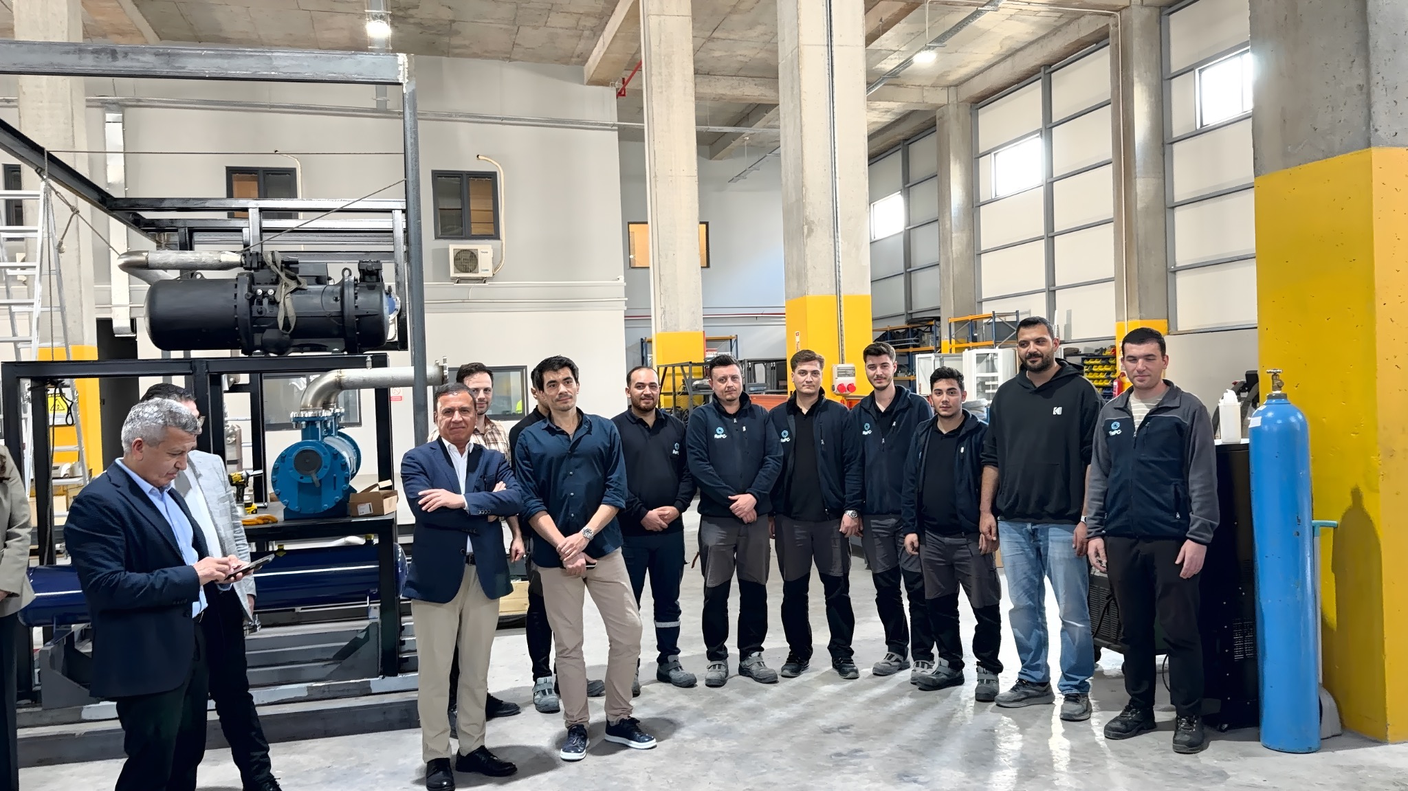 The visiting delegation and engineering team beside an equipment assembly in the workshop.