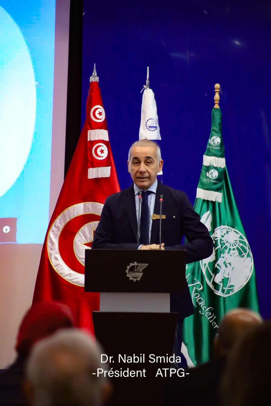 A speaker addressing the forum from the lectern, with the event flags behind him